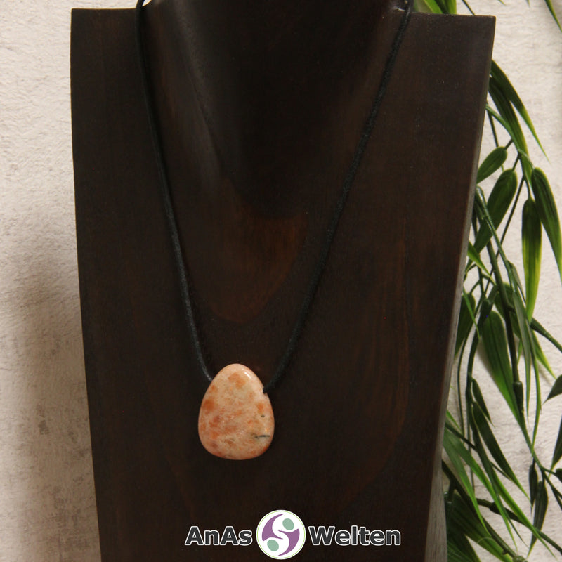 Ein gebohrter Sonnenstein Trommelstein an einem Baumwollband. Der Edelstein hat eine orange-beige Farbe, deren Muster ein wenig an Flammen erinnert.