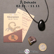 Die Sternzeichenkarte und je ein beispielhafter gebohrter Obsidian Trommelstein am Band und ein Fluorit Trommelstein der 2. Dekade liegen nebeneinander.
