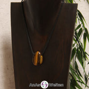 Ein gebohrter Tigerauge Trommelstein an einem Baumwollband. Der Edelstein hat eine hell- bis dunkelbraune Farbe, die je nach Lichteinfall wechseln kann und golden schimmert.