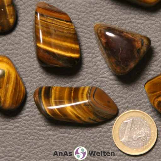 Das Bild zeigt einen Tigerauge Trommelstein anhand mehrerer Beispielsteine. Die Edelsteine sind eine Mischung aus dunkelbraun, hellbraun und gelb-golden. Die Farbe ändert sich dabei je nach Lichteinfall.