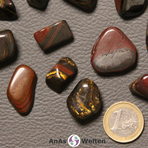 Das Bild zeigt einen Tigereisen Trommelstein anhand mehrerer Beispielsteine. Die Edelsteine haben fast alle eine rostrote, braune oder schwarze Grundfarbe. Sie sind oft gestreift und einige von ihnen weisen einen gold-gelben Glanz auf.